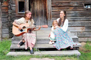Nainen soittaa kitaraa puutalon portailla ja toine nnainen pitelee tyttöä vierellään ja kuuntelee kitaransoittajaa
