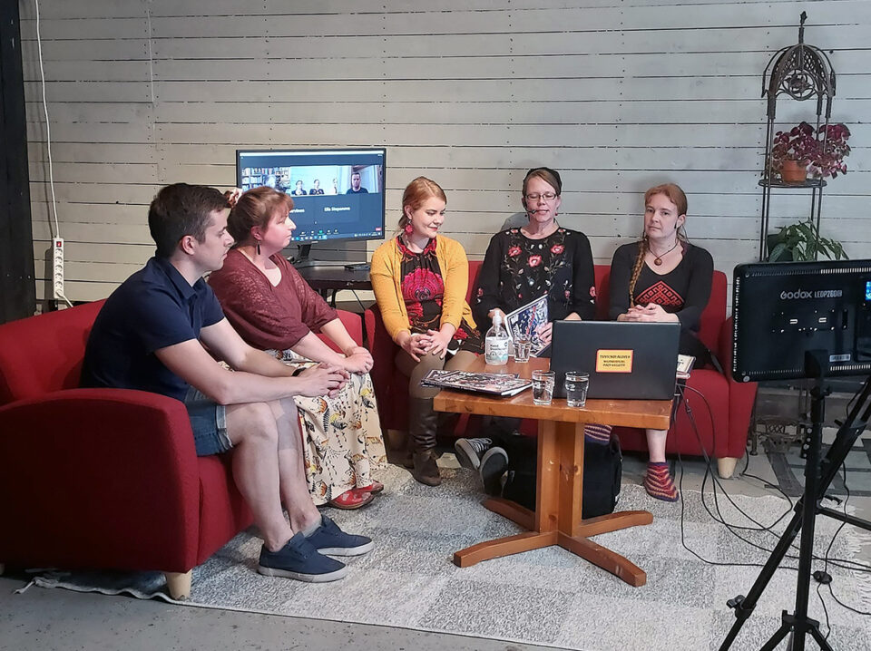 Neljä naista ja yksi mies istuvat punaisilla sohvilla, taustalla valkoinen tiiliseinä ja kamera kuvaa tätä keskustelua.