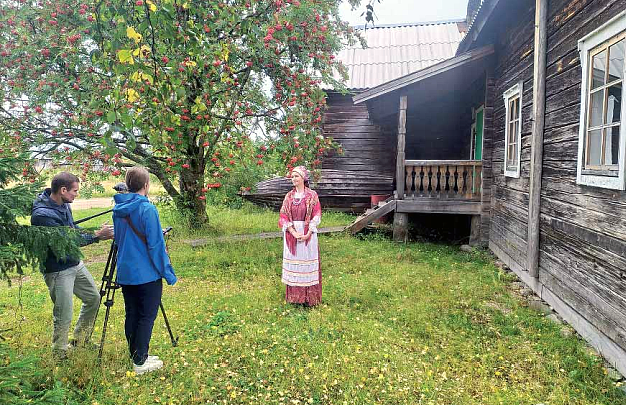 Nainen ja mies kuvaava karjalaisessa punaisessa perinneasussa esiintyvää naista harmaan puutalen edessä ja ympärillä on vihreää ja vehreää luontoa