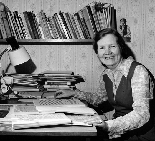 Hymyilevä nainen pöydän äärellä, jossa on paljon kirjoja ja seinällä on kirjahylly.