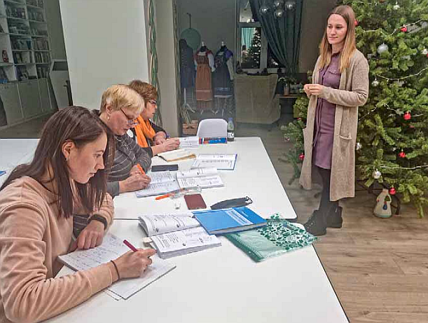 Nainen joulukuusen edessä tarkkailee kun ryhmä opiskelijoita kirjoittaa pöydän äärellä