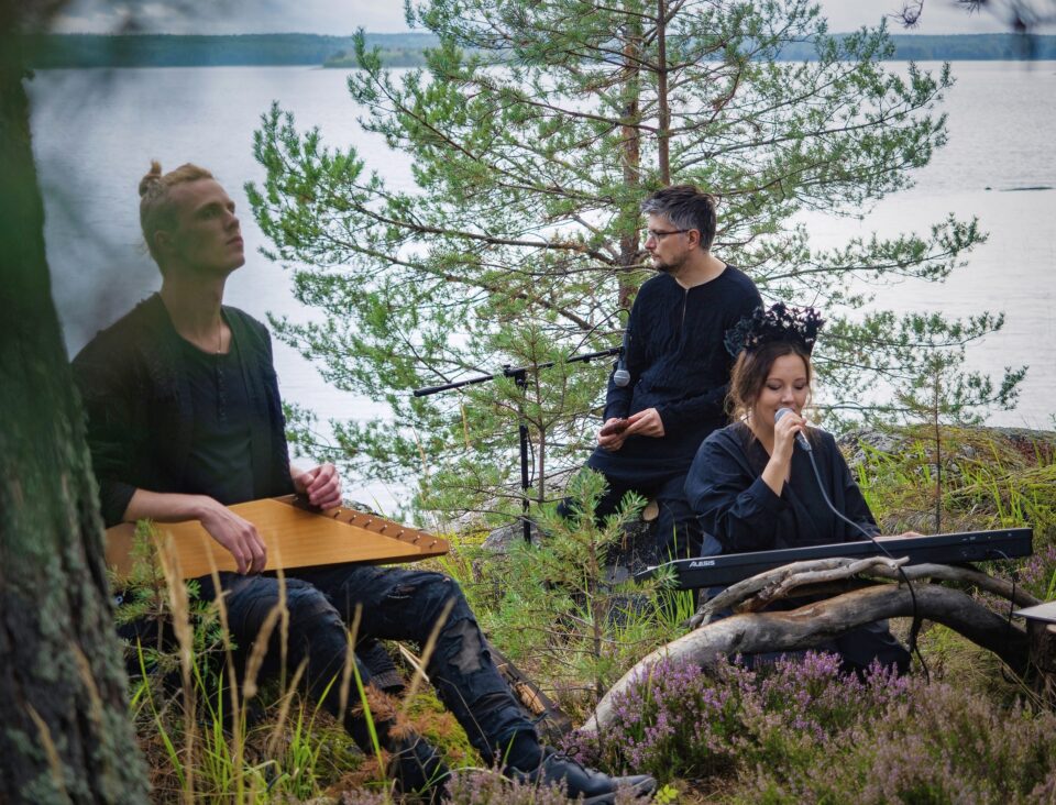 Kaksi miestä ja yksi nainen metsässä. Mies soittaa kanteletta, nainen soittaa koskettimia ja laulaa.