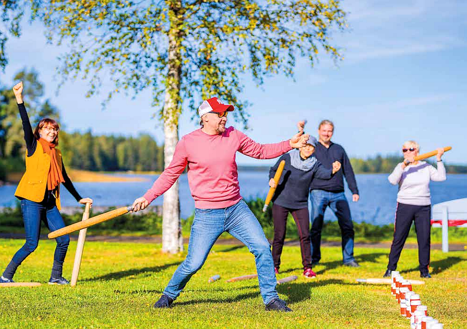 Kolme naista ja kaksi miestä nurmikolla kilpailemassa, taustalla näkyy sininen järvi