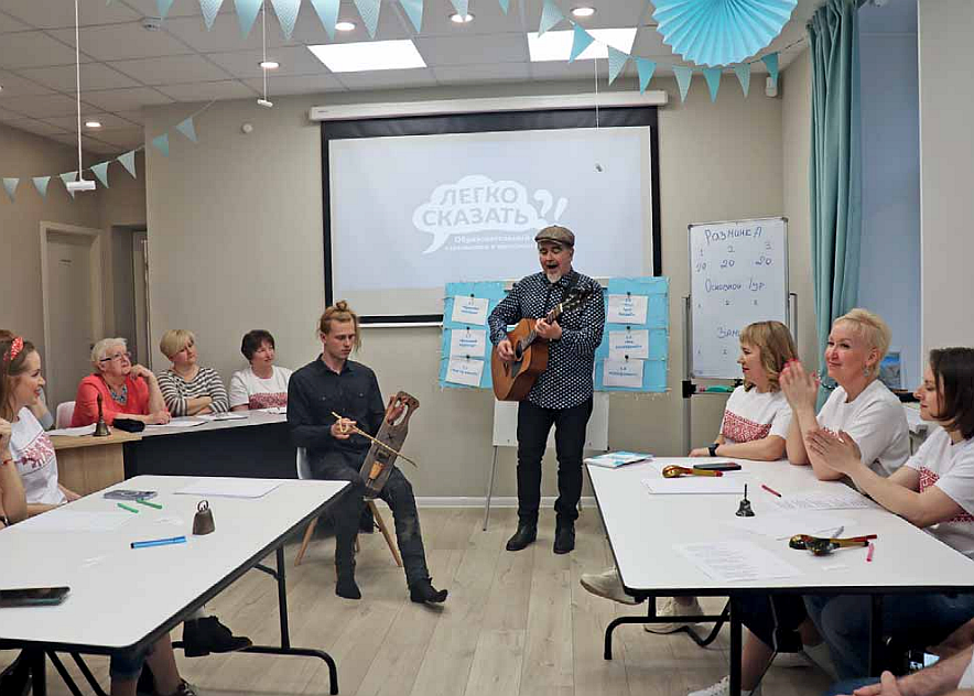 Mies esiintyy kitaran kanssa luokkahuoneessa ja muut ihmiset seuraavat istuen pöytien äärellä.