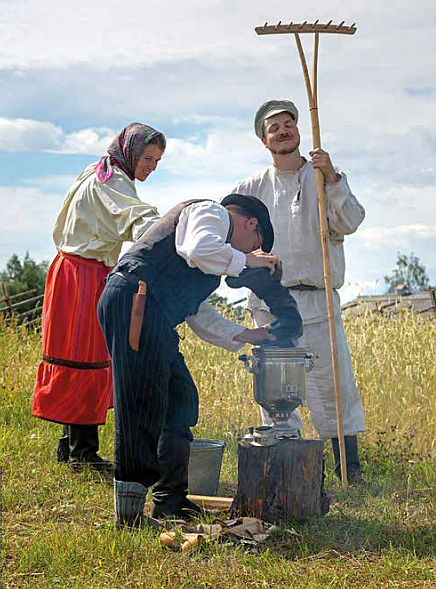 kaksi miestä ja yksin nainen pellolla heinätöissä, toinen mies pitelee haravaa pystyssä ja toinen täyttää samovaaria