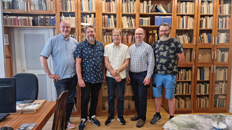 Viisi miestä seisoo suuren kirjahyllyn edessä.