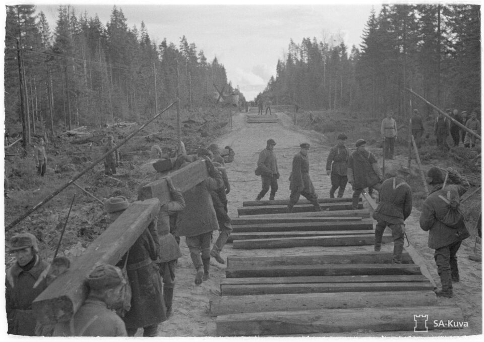 Rautatietöitä, miehet kantavat rautateiden palkkeja paikoilleen metsän keskellä mustavalkoisessa kuvassa