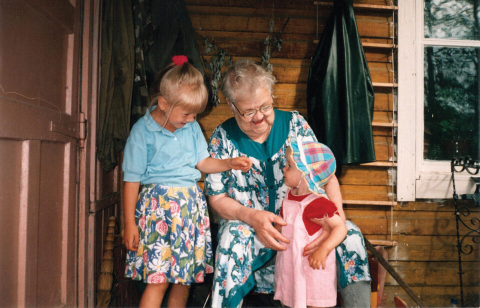 Vanha nainen istuu oven vierellä kaksi lapsenlapsenlastaan vierellään.