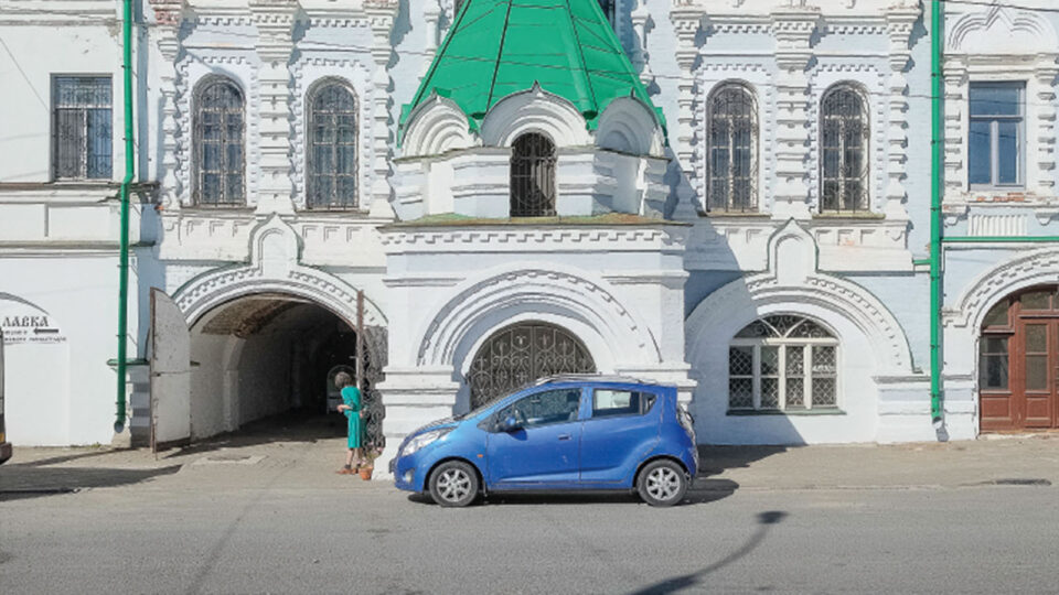 Henkilöauto suuren rakennuksen edessä, nainen kurkistaa porttikongiin.