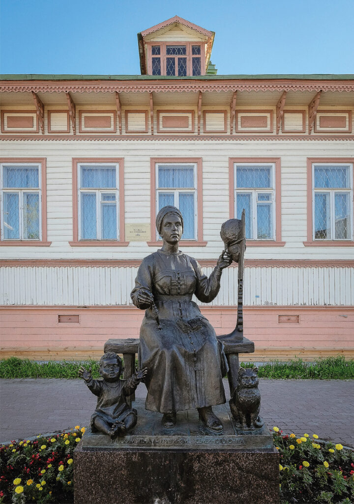 Äiti-lapsi-kissa -patsas, ympärillä kukkaistutuksia, takana puutalo.