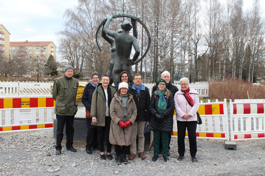 Yhdeksän ihmistä on asettunut ryhmäkuvaan patsaan juurelle puistossa.