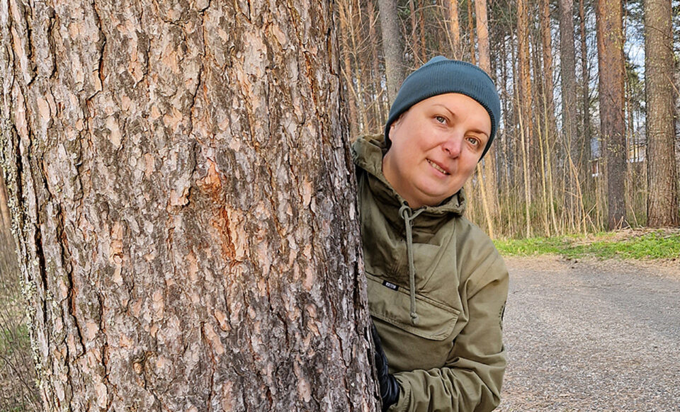Pipopäinen nainen kurkkaa suuren männynrungon takaa.