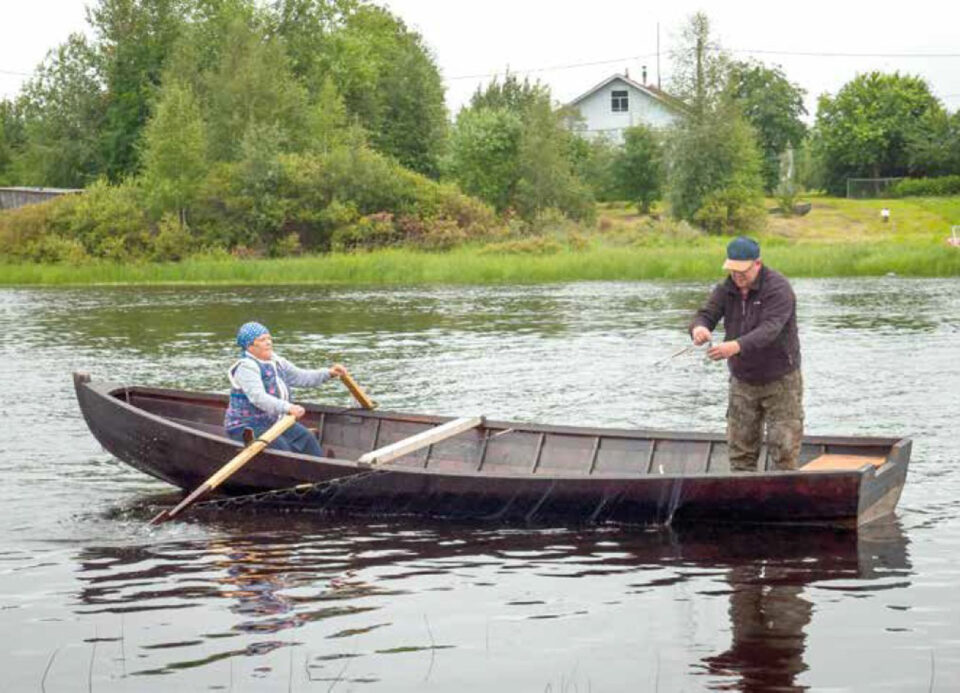 Puinen tummanruskea soutuvene kesäisellä järvellä, lippalakkipäinen mies seisoo veneen perässä verkkoja kokemassa, huivipäinen nainen istuu veneen keulassa käsissään airot, valmiina soutamaan tai huopaamaan.