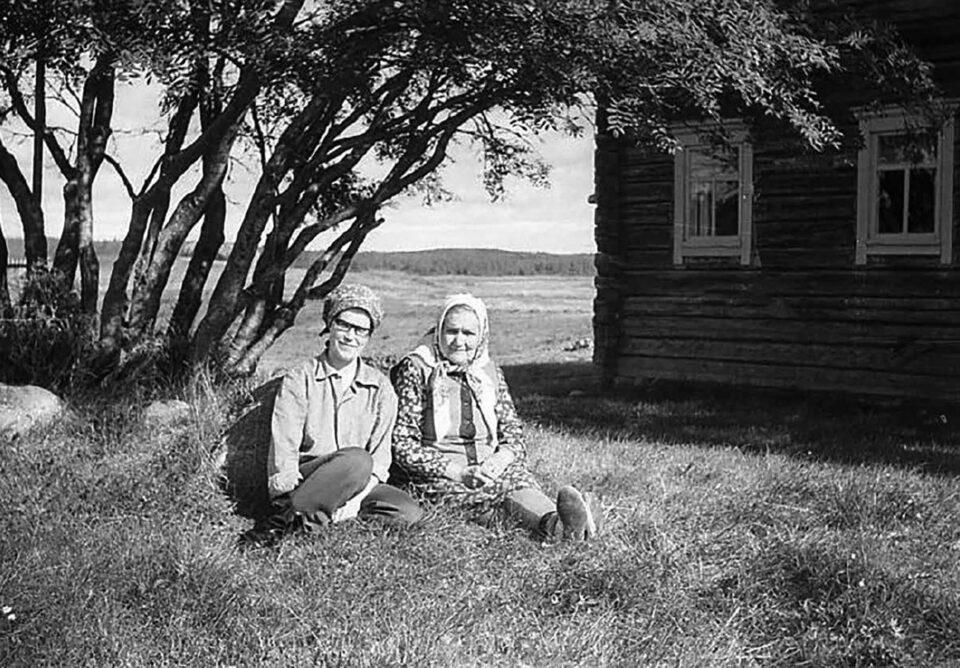 Mustavalkokuvassa kaksi huivipäistä naista istuu heinikossa puun juurella aurinkoisena kesäpäivänä, oikealla näkyy hirsitalon nurkka.