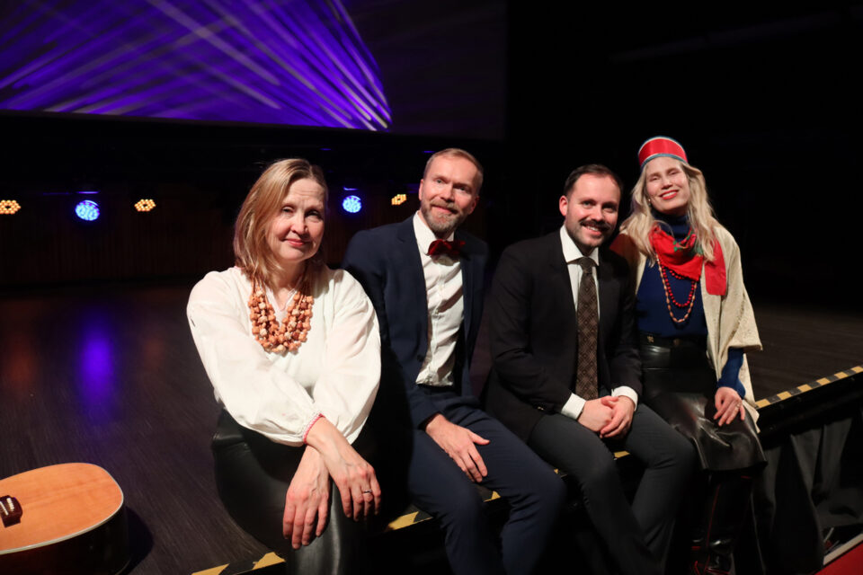 Neljä juhlavasti pukeutunutta ihmistä, kaksi naista ja kaksi miestä, istuvat esiintymislavan etureunalla katsoen iloisesti hymyillen kameraan.