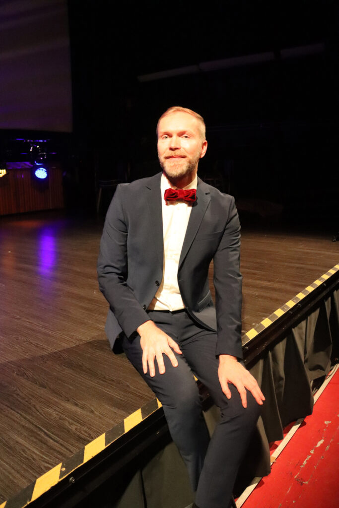 Tummaan pukuun ja tummanpunaiseen rusettiin pukeutunut mies, Jyrki Haapala, istuu esiintymislavan reunalla ja hymyilee kohti kameraa.