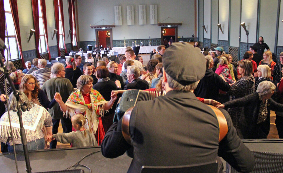 Etualalla harmonikansoittaja, juhlasalissa selin kameraan, hänen edessään iso joukko ihmisiä tanssii käsi kädessä letkana.