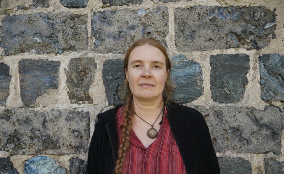 nainen katsoo suoraan kohti kameraa, taustalla jykevä harmaa kiviseinä.