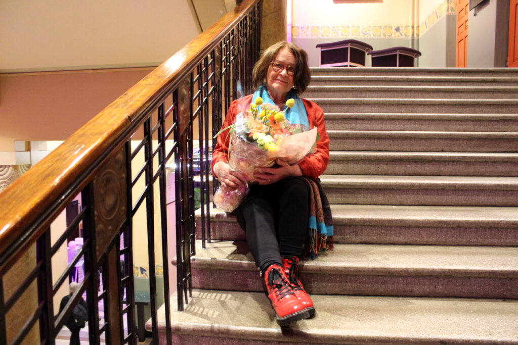 Juhlavasti pukeutunut nainen istuu arvotalon sisäpuolen portaikossa suuri, värikäs kukkakimppu sylissään ja katsoo kameraan.