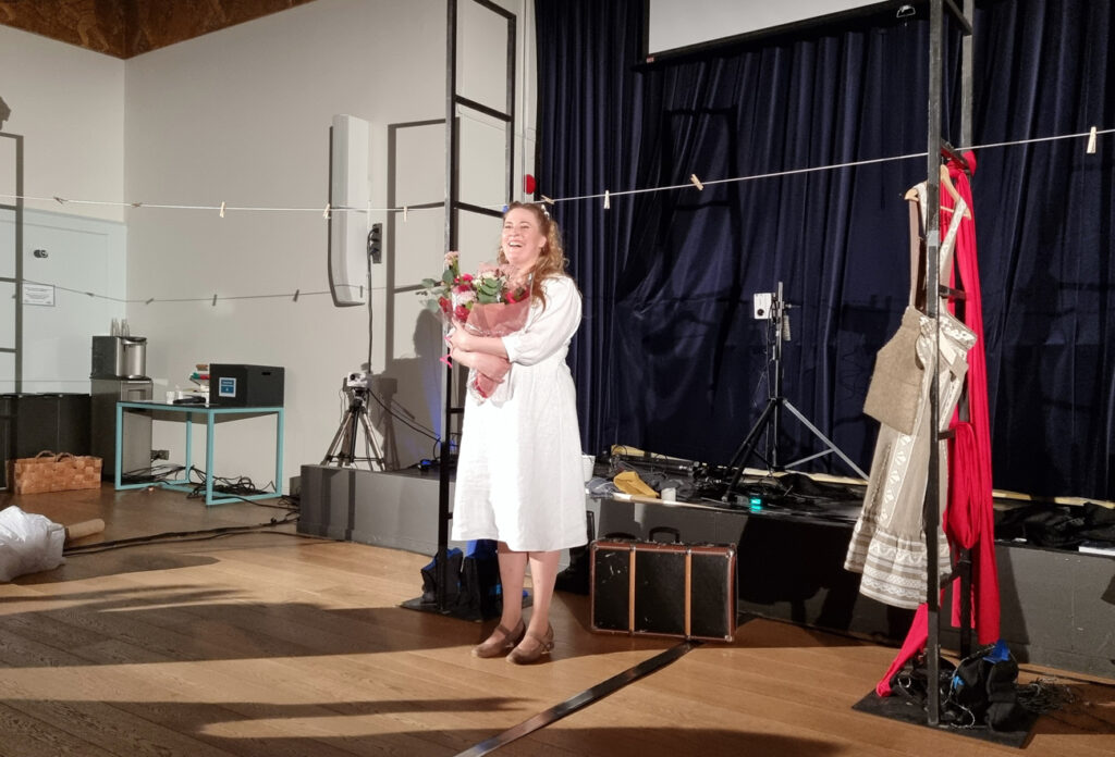 Teatterilavalla seisoo onnellisesti hymyilevä nainen valkoisessa leningissään, sylissä hyvin suuri ja värikäs kukkakimppu.