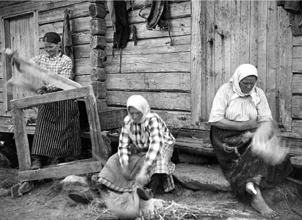 Hirsitalon edustalla ulkona kolme huivipäistä maalaisnaista käsittelee villoja, jokaisella on oma askareensa menossa. Mustavalkoisesta kuvasta voi päätellä, että se on otettu ehkä 1800-luvun puolella.