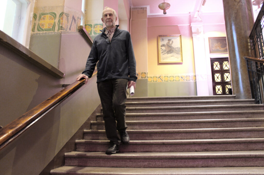 Iäkäs mies laskeutuu leveitä portaita jugendtyylisen talon sisätilassa. 