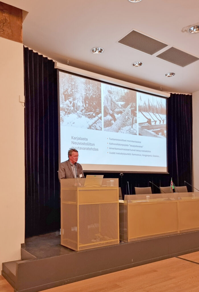 Mies puhuu mikrofoniin seminaarisalin puhujankorokkeella, taustalla suuri valkokangas, jossa esitysdiat ovat näkyvissä.