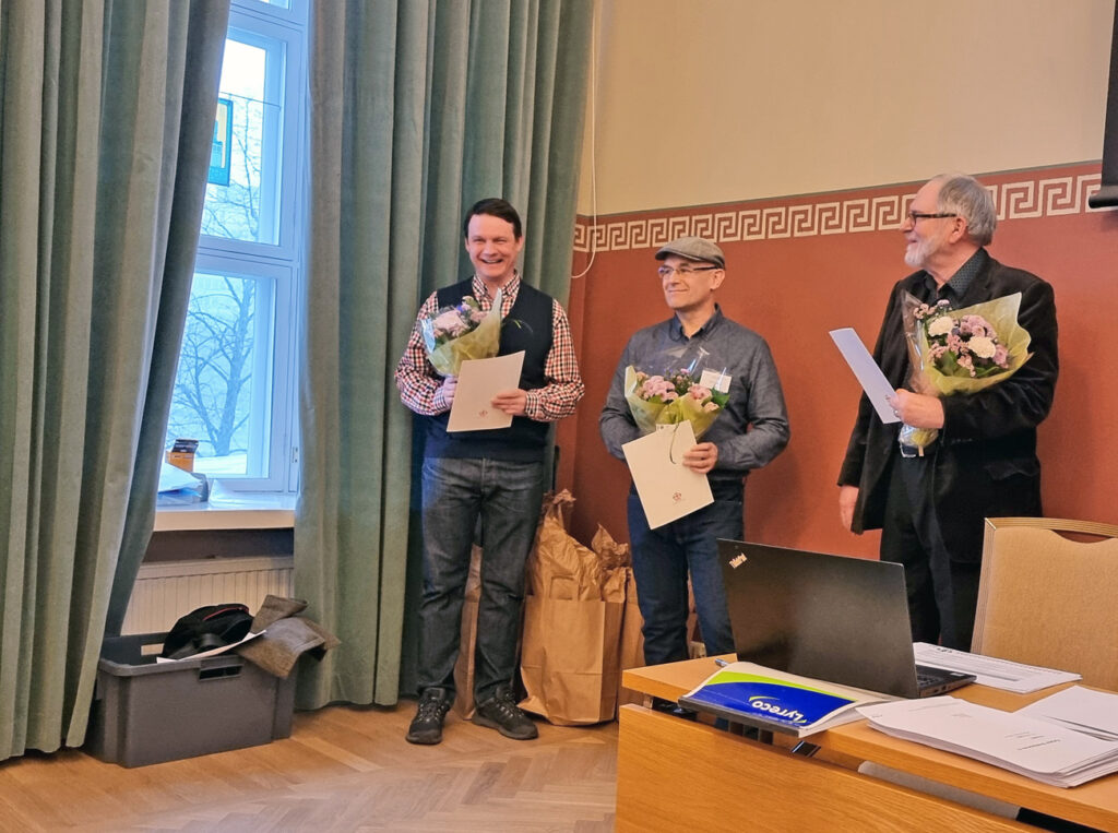 Kolme miestä hymyilee sylissään suuret kukkakimput ja kädessään kunniakirja.