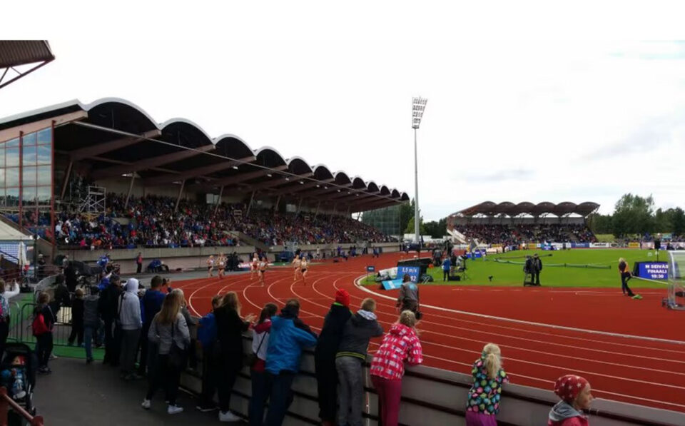 Urheilustadionin katsomossa runsaasti yleisöä, radalla kilpajuoksijoita ja keskialueella muutamia toimitsijoita.