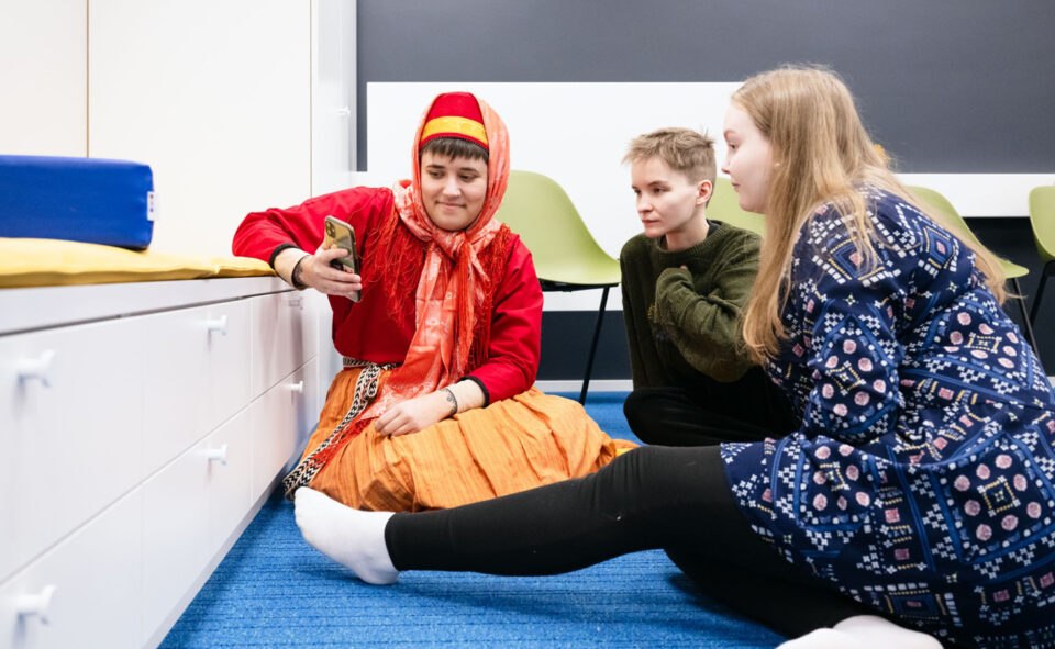 Kolme nuorta aikuista istuu lattialla keskustellen, yksi heistä on pukeutunut värikkäästi karjalaisnaisen perinneasuun, kaksi muuta on arkivaatteissa.