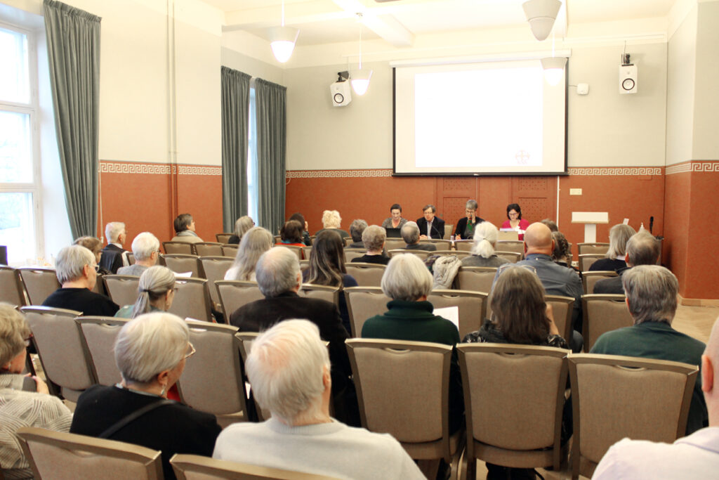 Kokoussalissa on kymmeniä ihmisiä selin kameraan, heidän edessään istuu neljä henkilöä rivissä, pitkän pöydän ääressä kasvot kohti yleisöä.