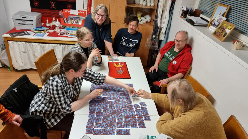 Ryhmä ihmisiä on pöydän ääressä, kumartuneena pelaamaan värikästä muistipeliä.