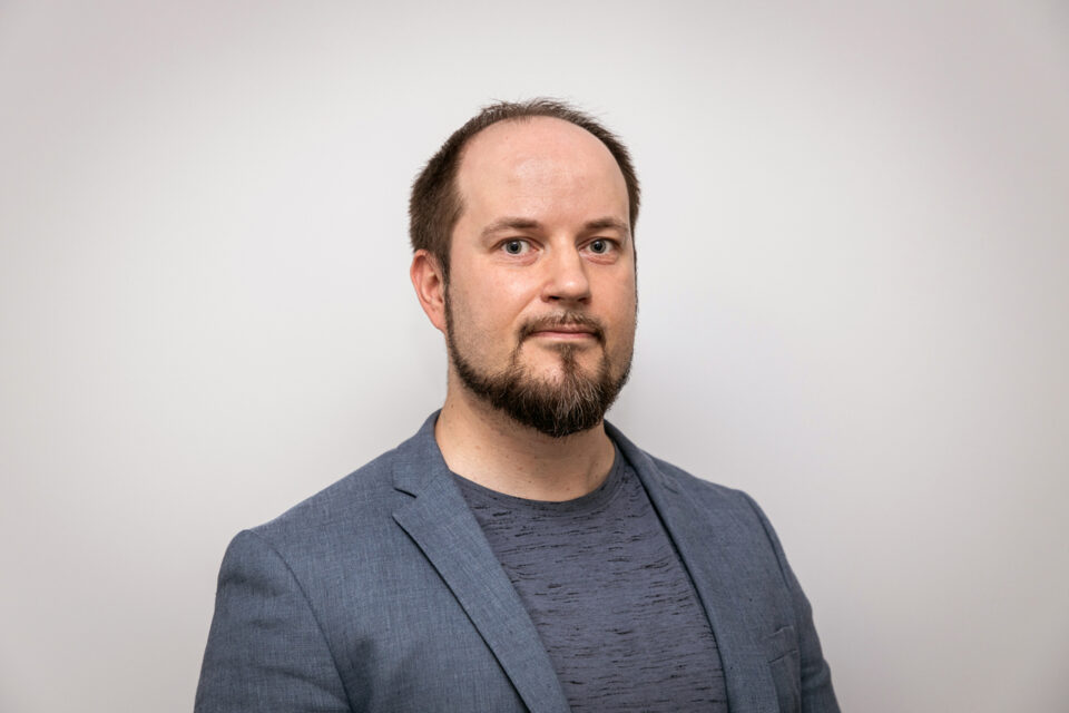 Puolilähikuva, jossa mies katsoo suoraan kameraan harmaa puvuntakki yllään.