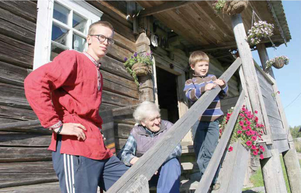 Vanhan hirsitalon edustalla, ulkoportailla aurinkoisessa kesäsäässä iäkäs mies, nuori mies ja leikki-ikäinen lapsi.