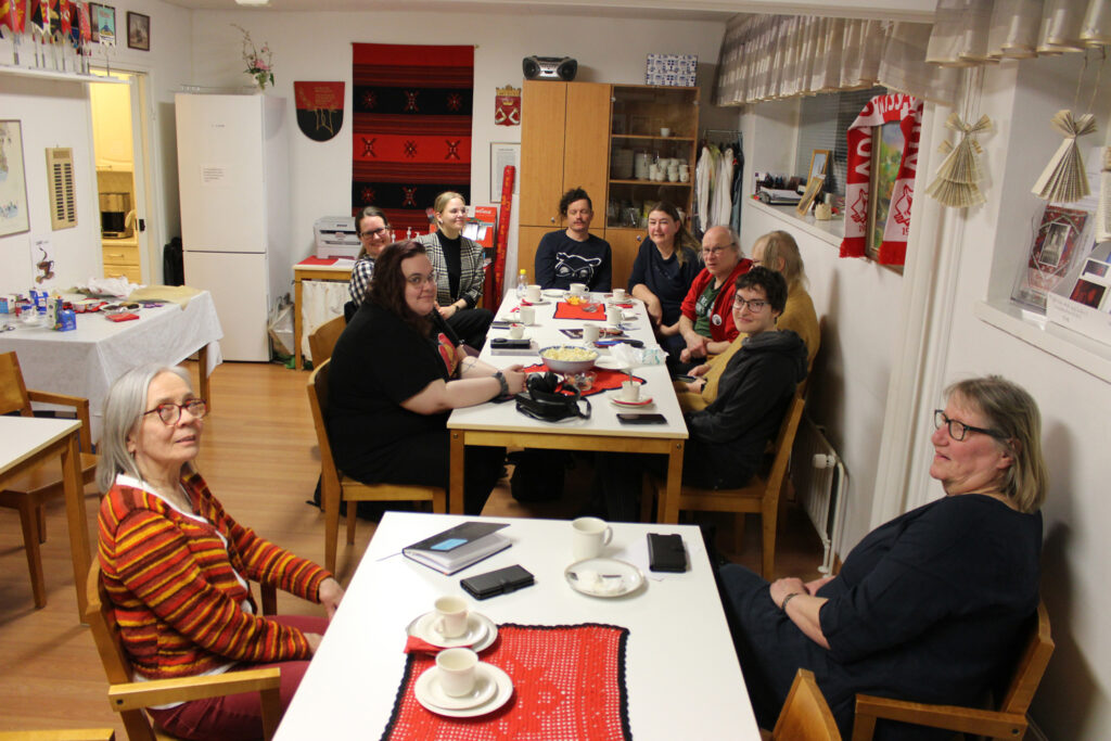 Ryhmä iloisia ihmisiä istuu kahden pitkän pöydän ääressä, takaseinällä on Karjala-värityksin koristettu seinävaate ja vaakuna, pöydillä punaiset kaitaliinat ja astioita kahvittelun jäljiltä.