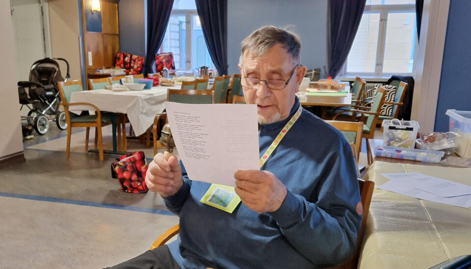 Iäkäs mies istuu lähikuvassa, lukee lukulasit päässään A4-kokoista paperia.
