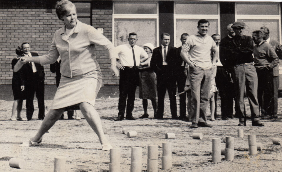 1960-luvun tyyliin, korkeaan kampaukseen ja vaaleaan kävelypukuun pukeutunut nainen heittää kyykkämailaa. Kravatteihin ja puvuntakkeihin sonnustautuneet miehet seuraavat intensiivisesti peliä kentän laidalla. Mustavalkokuva.