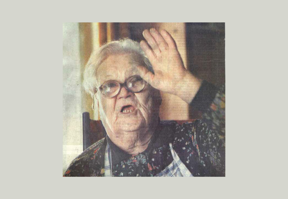 Iäkäs, silmälasipäinen nainen puhuu tai laulaa lähikuvassa ja kohottaa kättään ikään kuin tervehdykseen.