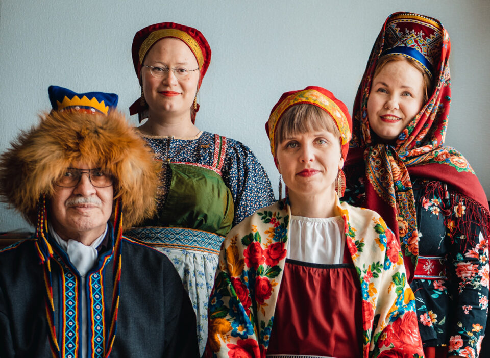 Karjalaisiin ja kolttasaamelaisiin perinneasuihin pukeutuneet kolme naista ja yksi mies hymyilevät kohti kameraa.
