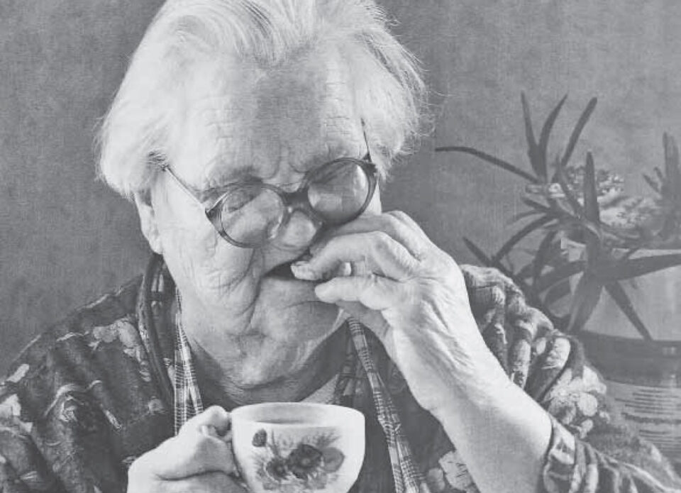 Mustavalkokuvassa iäkäs silmälasipäinen, valkotukkainen nainen kädessään kukallinen kahvikuppi, laittaa juuri suuhunsa kahvileipää.