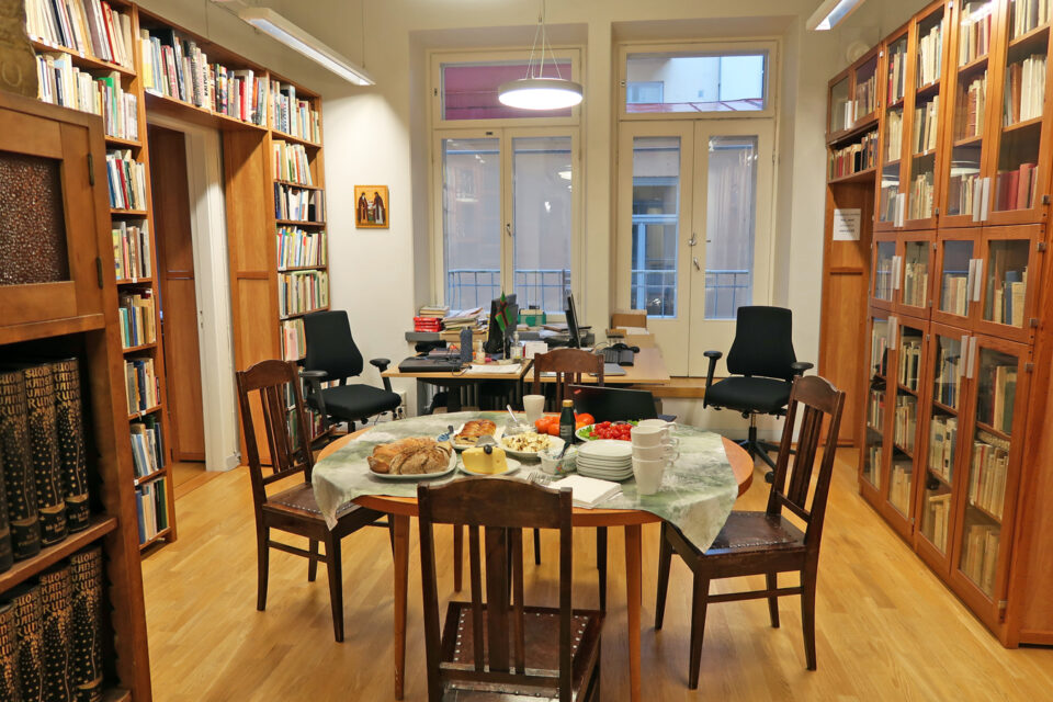 Suurten kirjahyllyjen reunustamassa huoneessa taustalla korkeat ikkunat, joiden edessä kaksi työpistettä. Edustalla pyöreä pöytä ja neljä tuolia sen ympärillä.