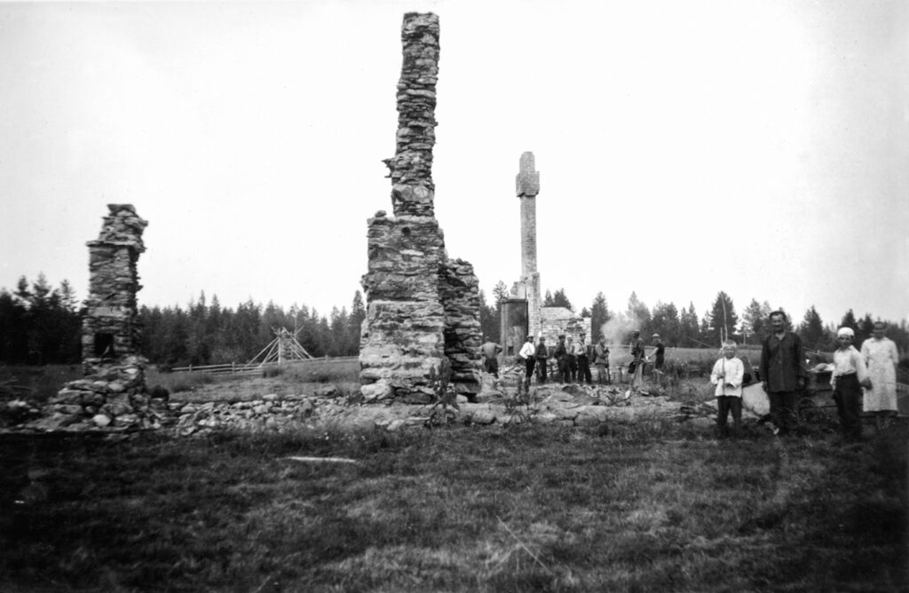 Vanhassa mustavalkokuvassa eri-ikäisiä ihmisiä seisoo poltetun kylän raunioilla, savupiiput kohoavat kohti taivasta ja kiviset perustukset lepäävät maassa, vaikka talot ovat kadonneet.