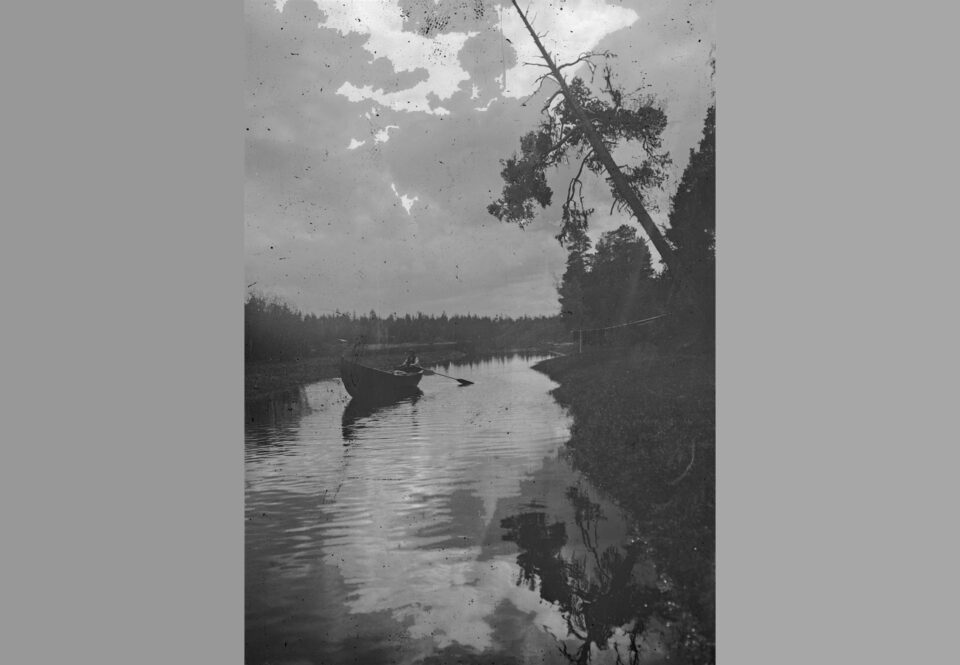 Vanhassa mustavalkoisessa valokuvassa kirkas, poutapilvinen taivas, jonka alla pitkä tyven joki, jolla puuveneellä soutaja.