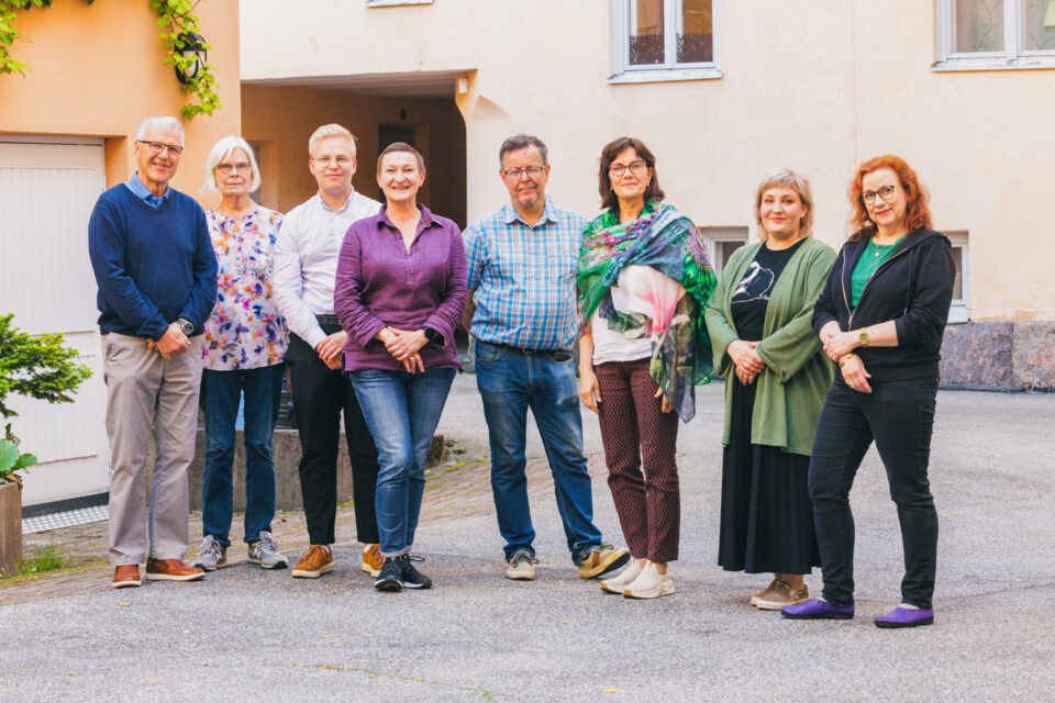 Kahdeksan hymyilevää ihmistä seisoo vanhojen kivitalojen ympäröimällä sisäpihalla. Kuvassa on vahva ja lämmin valo, ihmiset katsovat kohti kameraa.