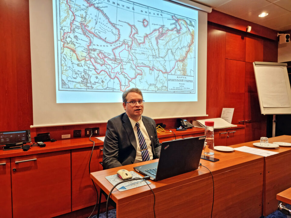 Mies istuu pöydän ääressä ja puhuu, hänen taakseen seinälle on heijastettu vanha suurikokoinen karttakuva Venäjältä, Arkangelin alueelta.