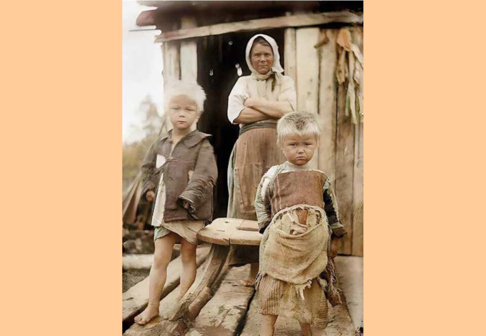 Kaksi resuista pikkulasta katsoo totisina kohti kameraa, taustalla huivipäinen nainen kädet puuskassa sekä lautahökkeli.