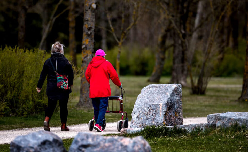 Nuorempi ja vanhempi nainen kävelevät puistotietä pitkin, kuvattuna takaapäin. Vanhemmalla punatakkisella naisella on rollaattori apuvälineenään.