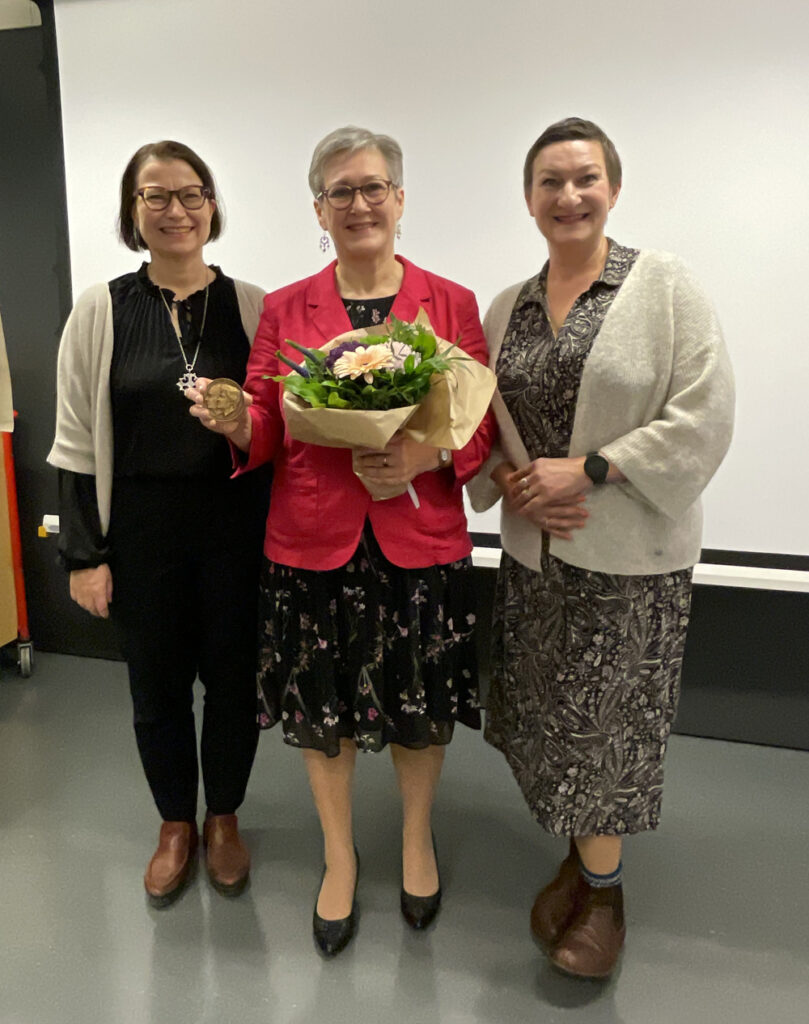 Kameraan katsoo kolme iloista naista, keskimmäisellä on kukkakimppu sylissään.