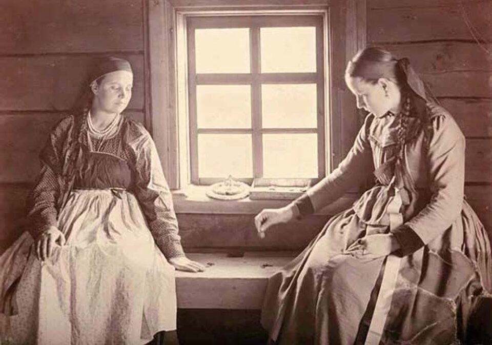 Seepiansävyisessä vanhassa valokuvassa kaksi nuorta tyttöä istuu penkillä, pieniruutuisen ikkunan edessä. Pelaavat noppaa.