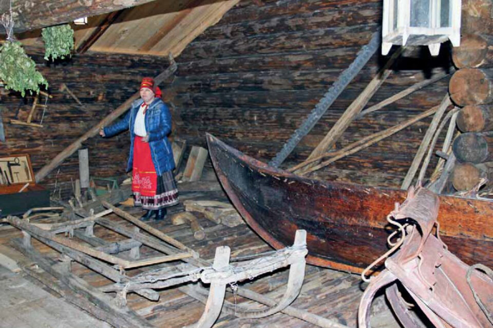 Värikkääseen vepsäläiseen kansanpukuun pukeutunut nainen seisoo tumman hirsirakenteisen huoneen takaosassa, edustalla vanhat reen jalakset ja puuvene.